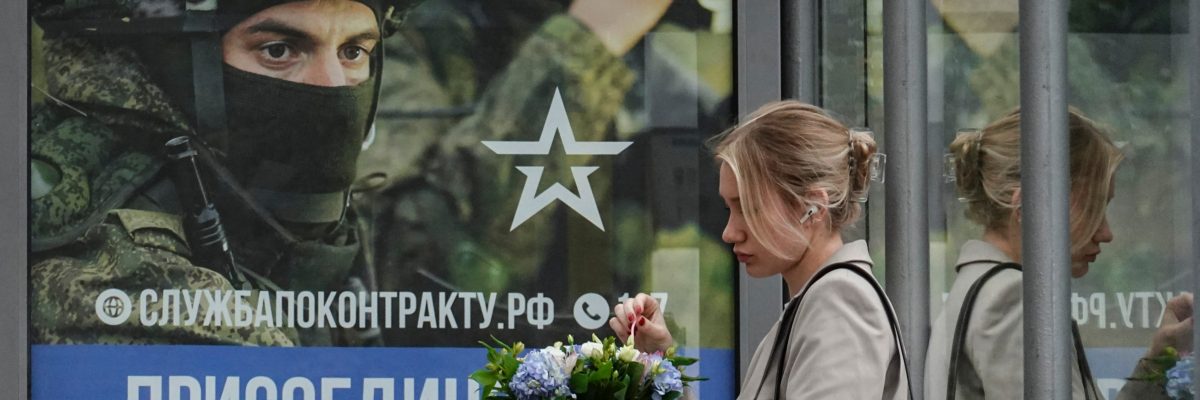 Žena čeká na autobusové zastávce v Moskvě, kde je plakát lákající muže do armády, 25. srpna 2023